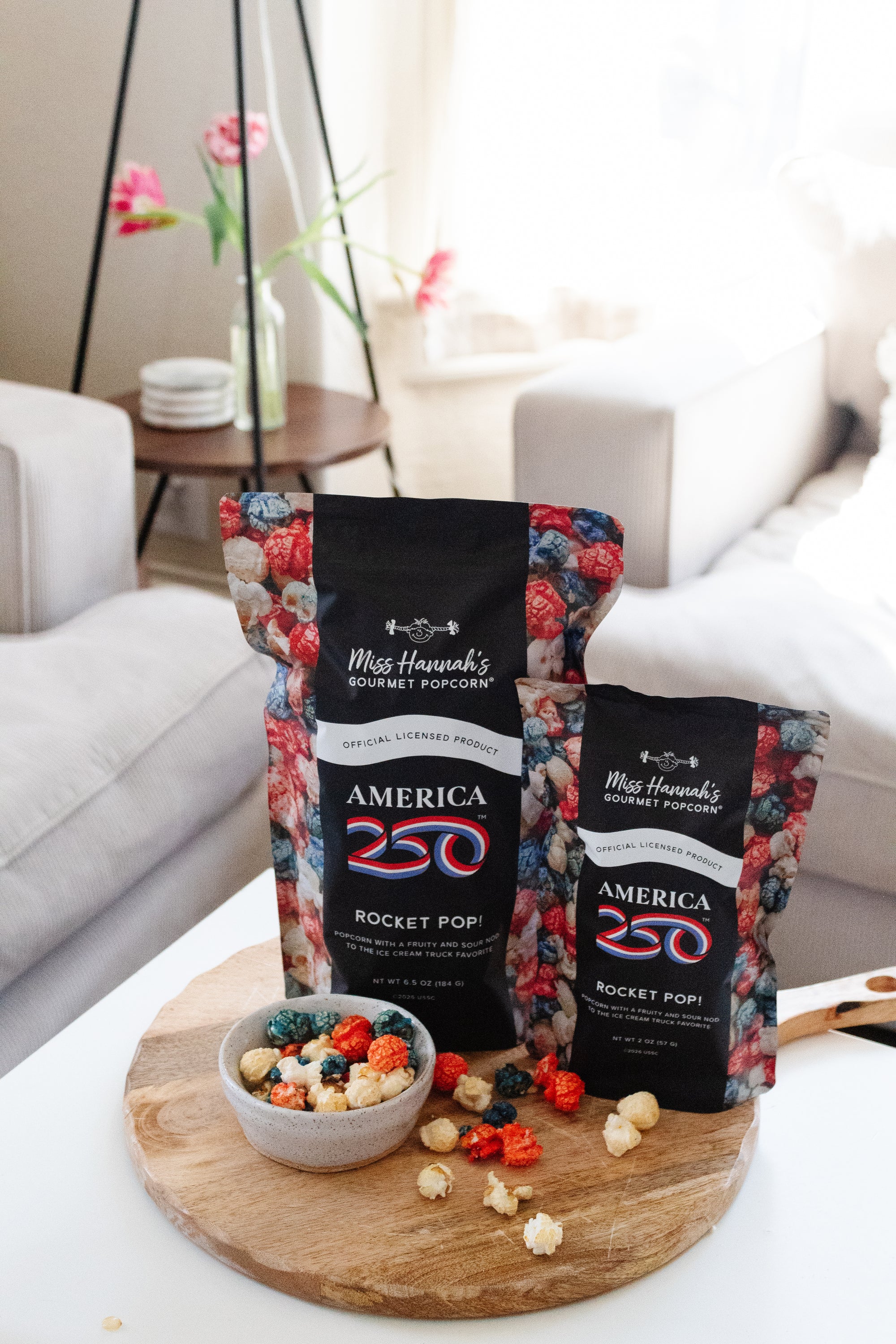 Two bags of Miss Hannah's Gourmet Popcorn on a wooden board with a bowl of popcorn in a living room setting.